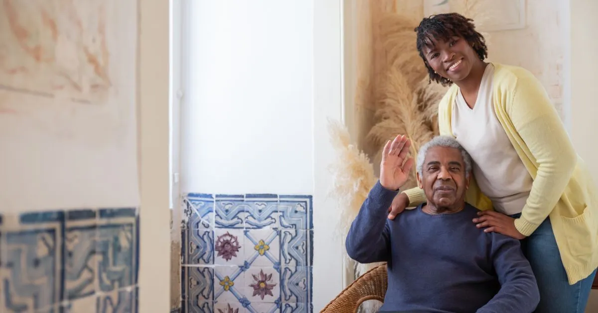 Adult daughter standing behind her elderly father at home, both smiling warmly