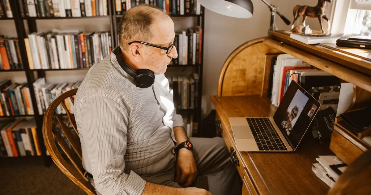Elderly man having a video call on his laptop at a home desk surrounded by books